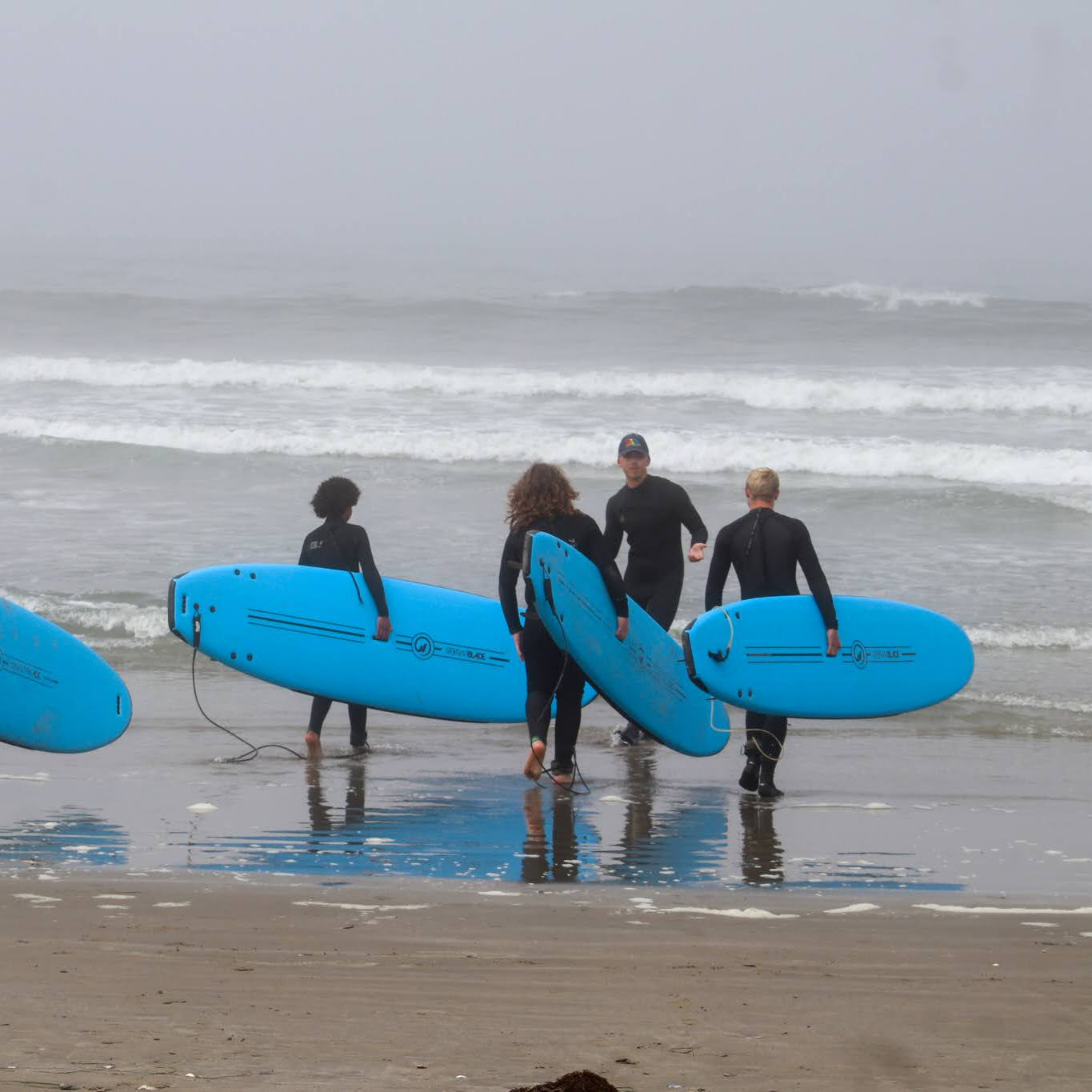 Kids and teens in wetsuits carrying blue surfboards along the beach at Camp Ocean Pines in California.
