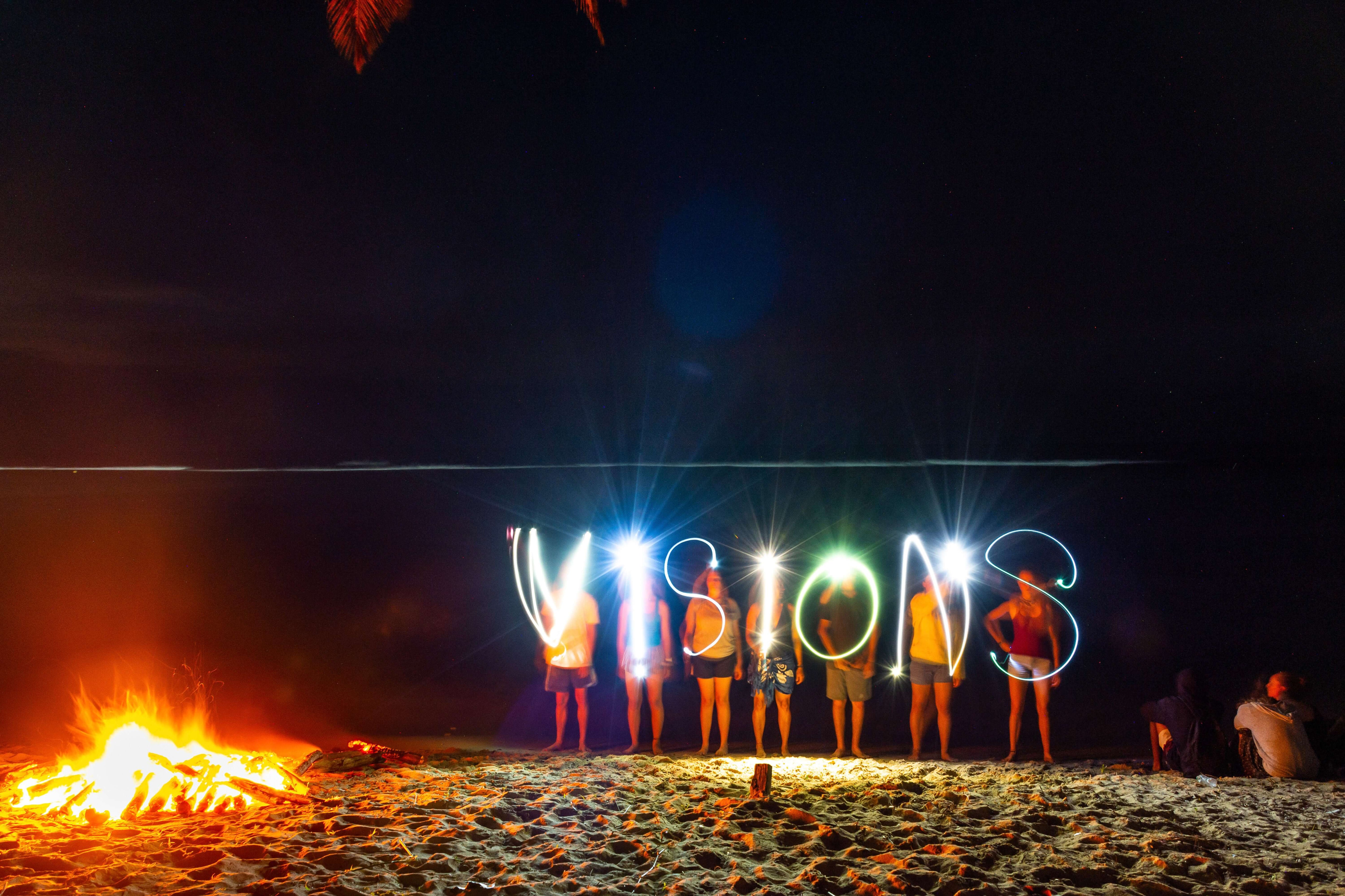 Group of people on beach at night, using lights to write "VISIONS" near a bonfire. Summer travel program atmosphere.