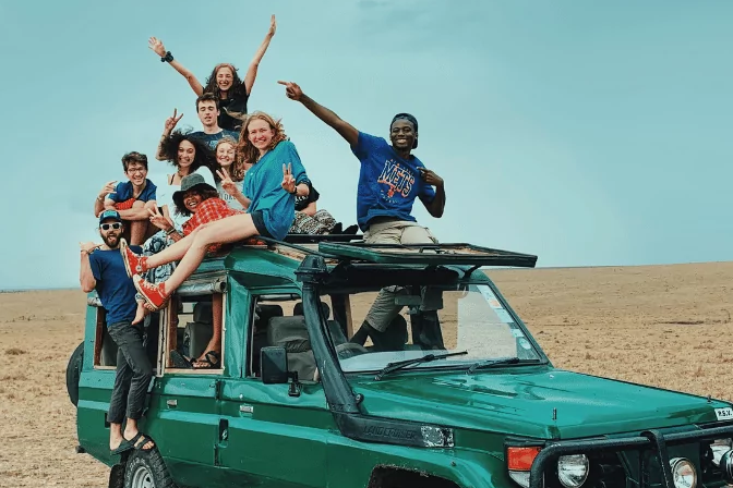Group of diverse teens joyfully posing on a green jeep during a summer camp adventure in an open field.