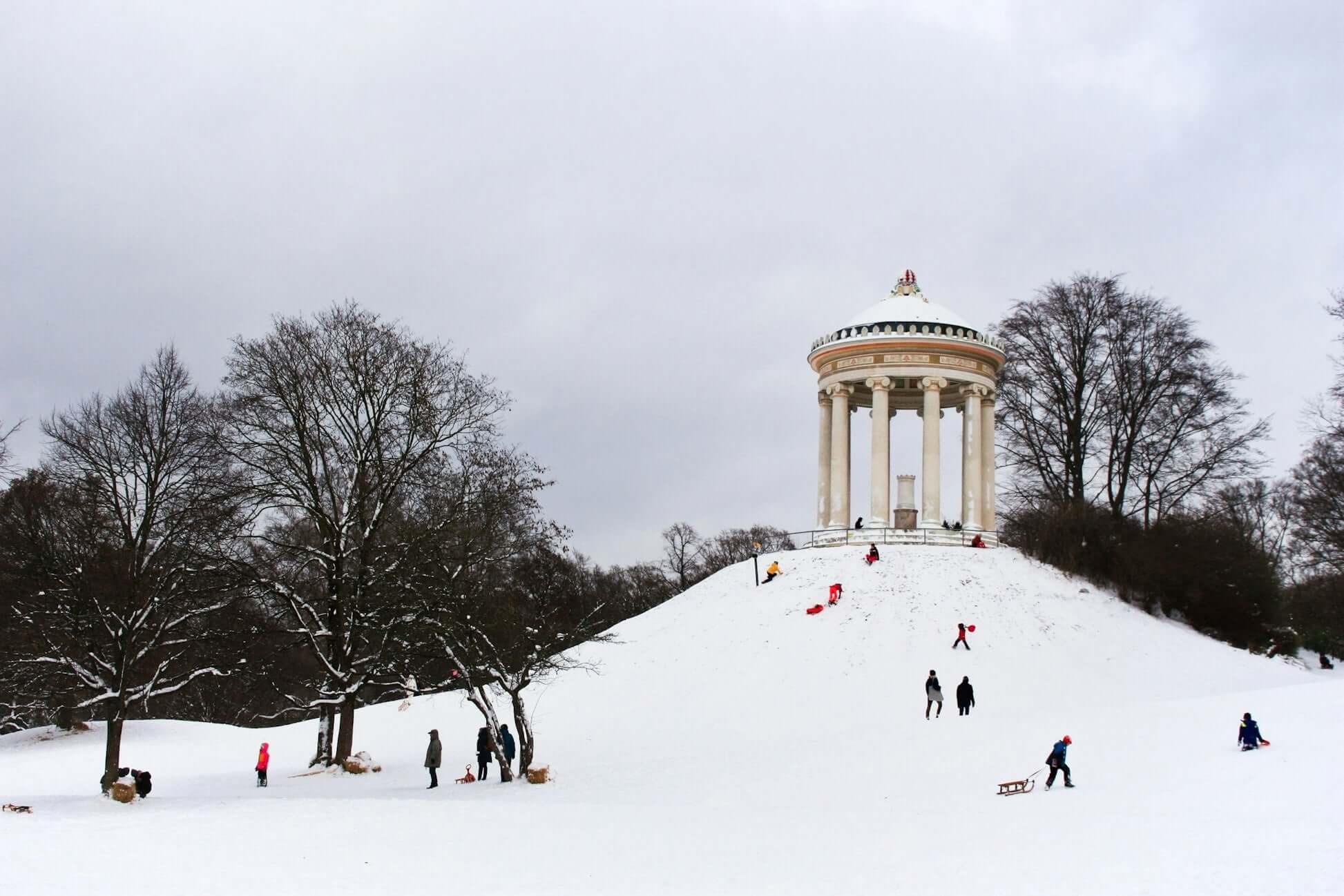 Snow-covered hill in Bavaria with children sledding and a historic pavilion in the background, depicting winter fun.