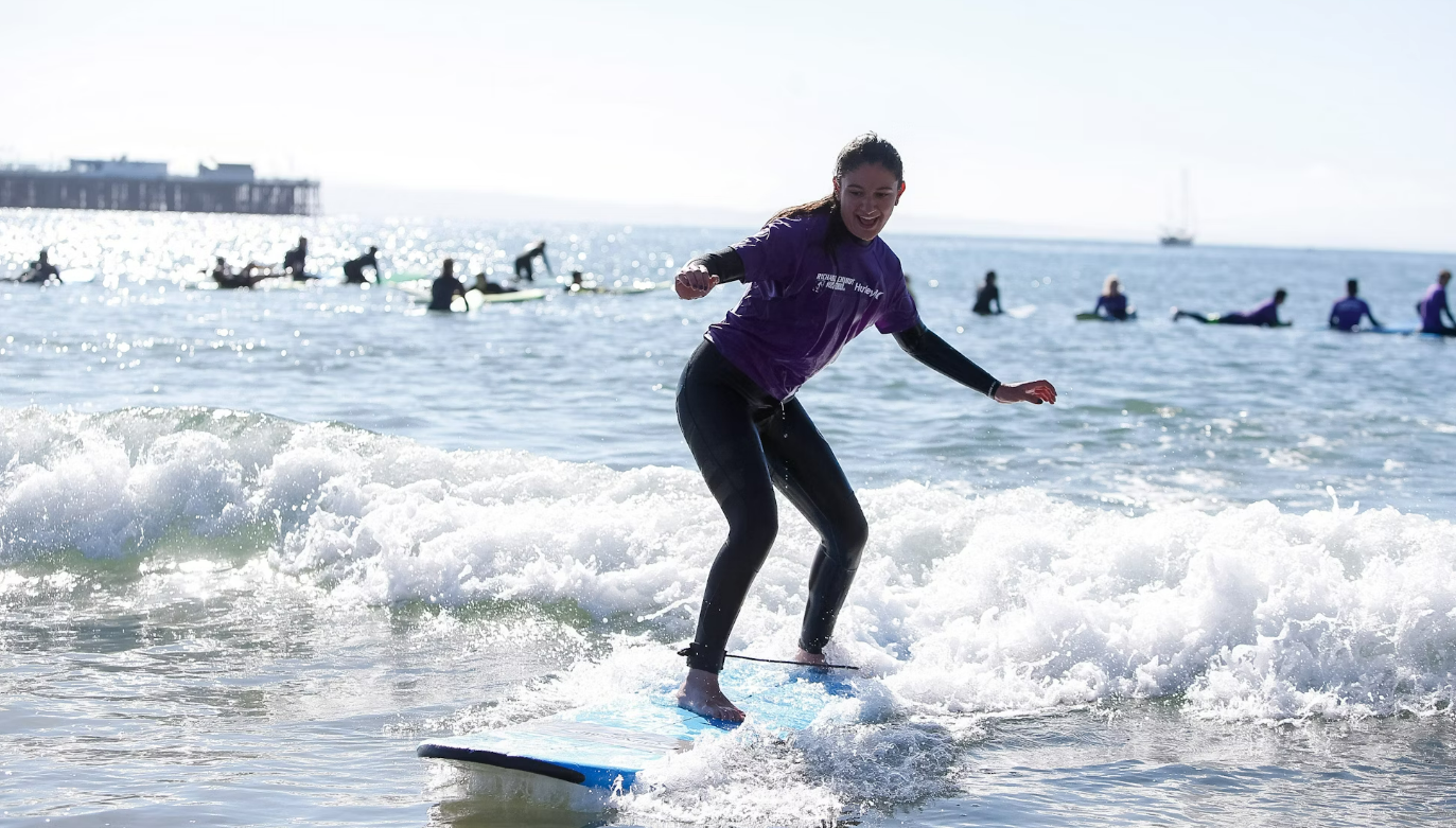 Teenager surfing in the ocean during a summer camp, enjoying summer programs at a beach with other participants in the background.