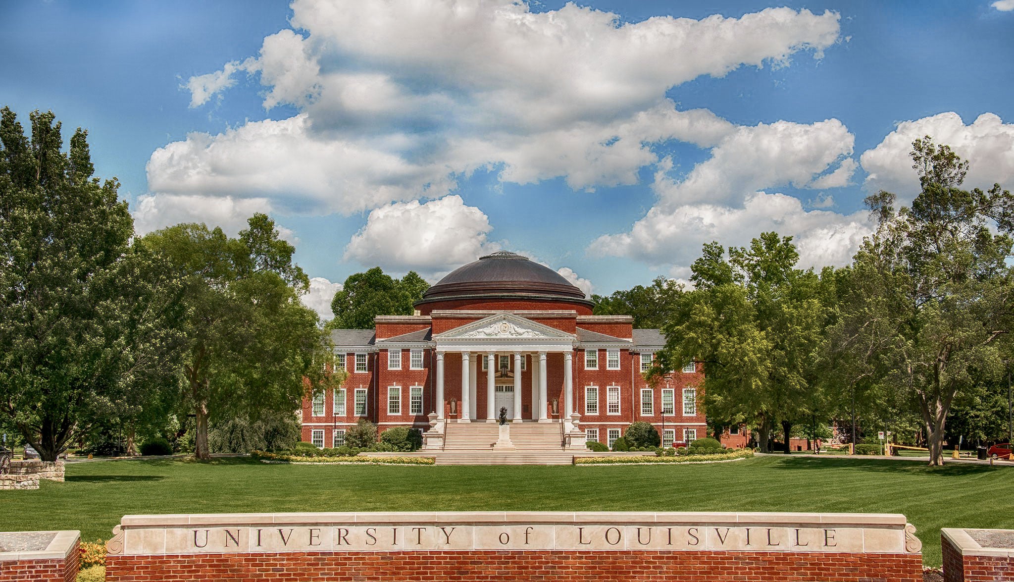 University of Louisville campus view featuring a historic building surrounded by lush greenery and a cloudy blue sky.