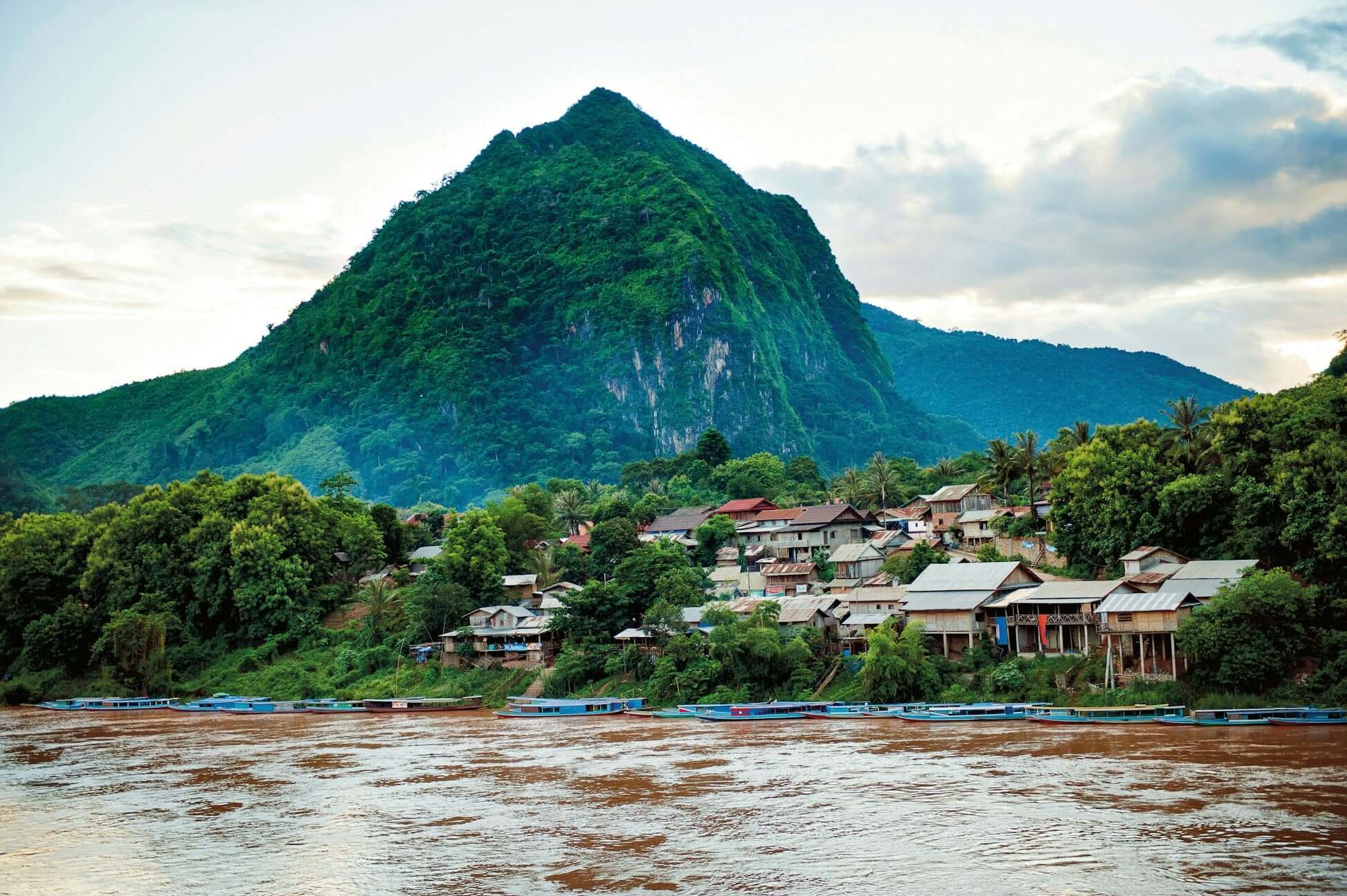 Scenic view of traditional houses along the Mekong River with a lush mountain backdrop in Thailand. Perfect for summer expeditions.