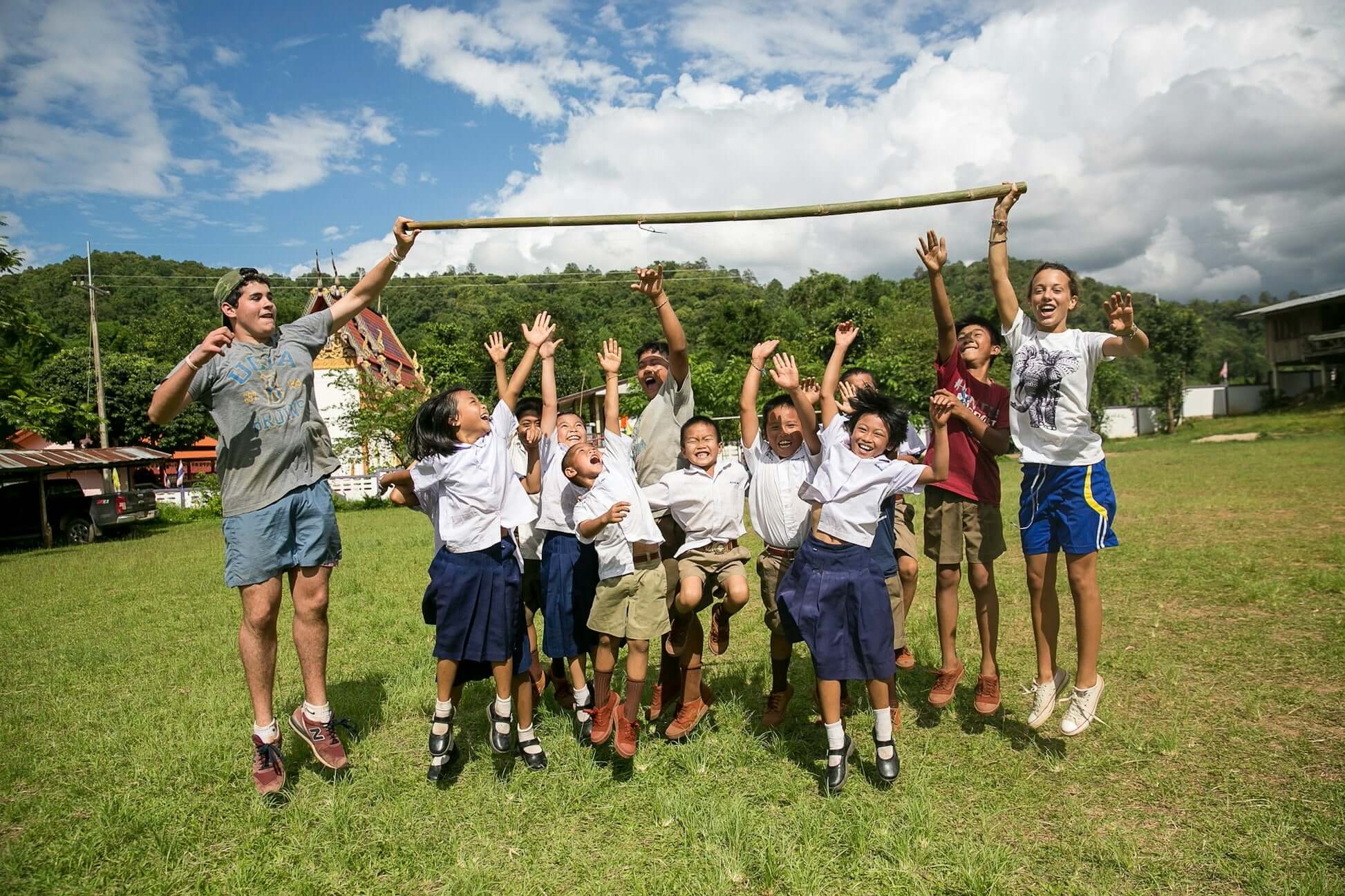 Teens enjoying a service expedition in Thailand, jumping with local children in a green landscape, promoting summer adventures.