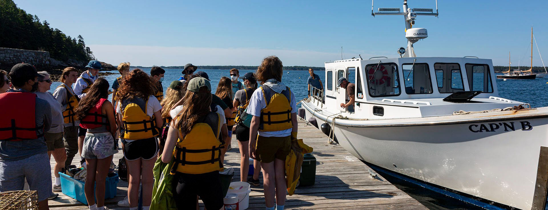 Group of high school and middle school students in life jackets boarding a boat for a summer camp program by the water.