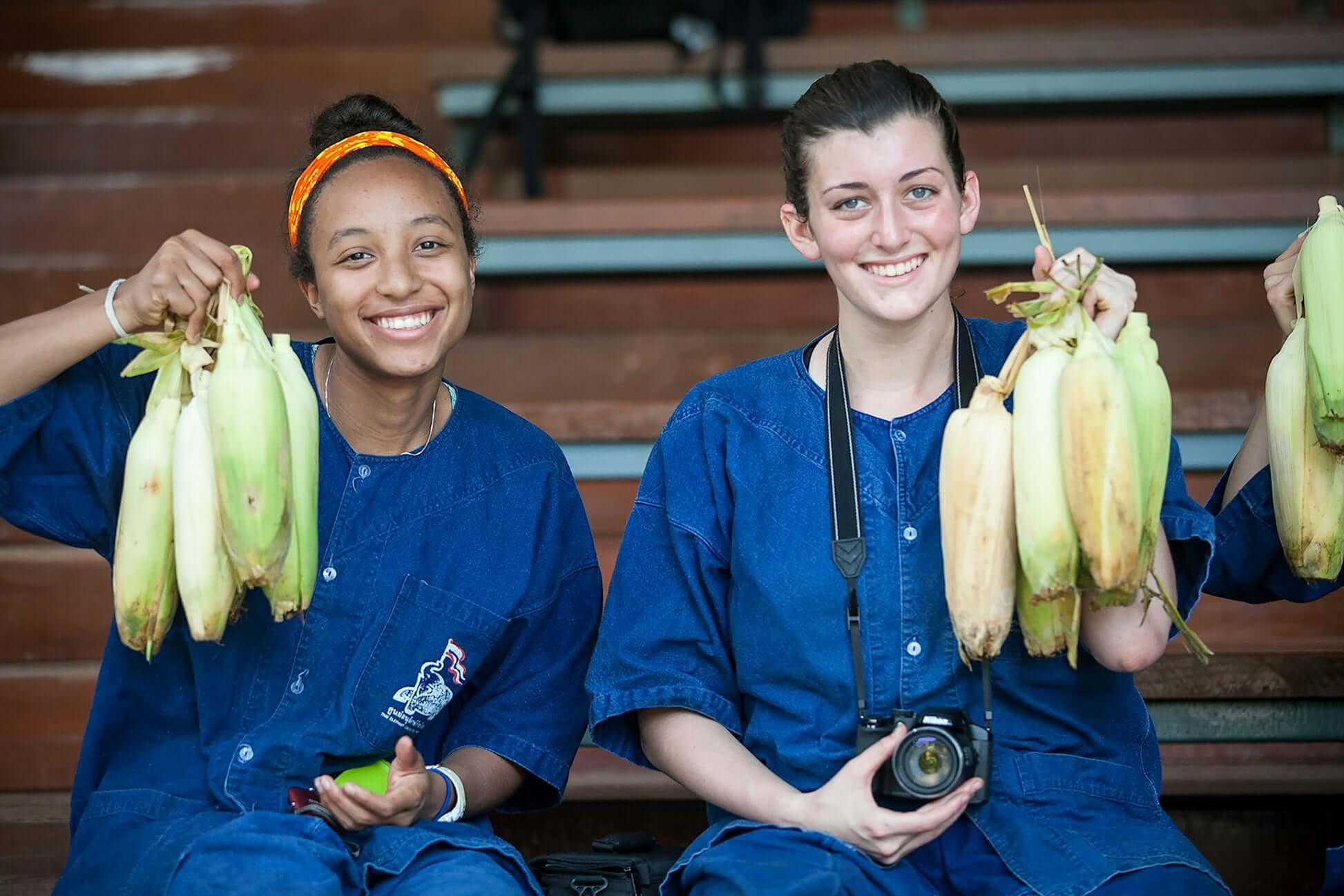 Two high school girls joyfully hold fresh corn, ready for the Thai Elephant Conservation Project adventure.