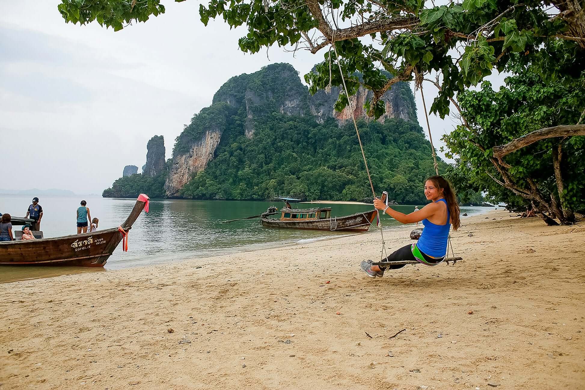 Woman enjoying a swing on a beach in southern Thailand with traditional boats and lush greenery in the background.