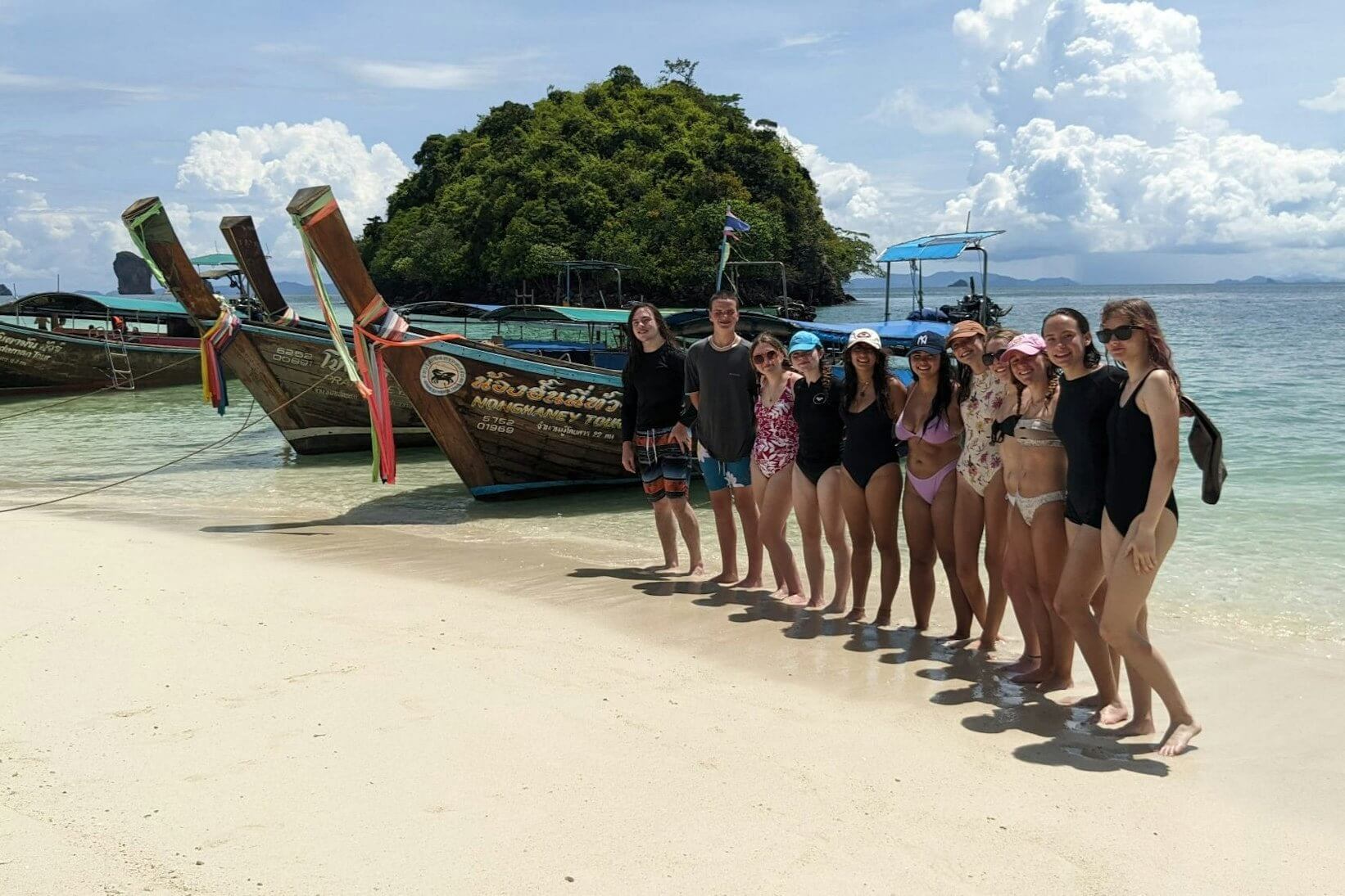 Group of students enjoying a beach in Thailand, standing by traditional boats during a summer travel experience.