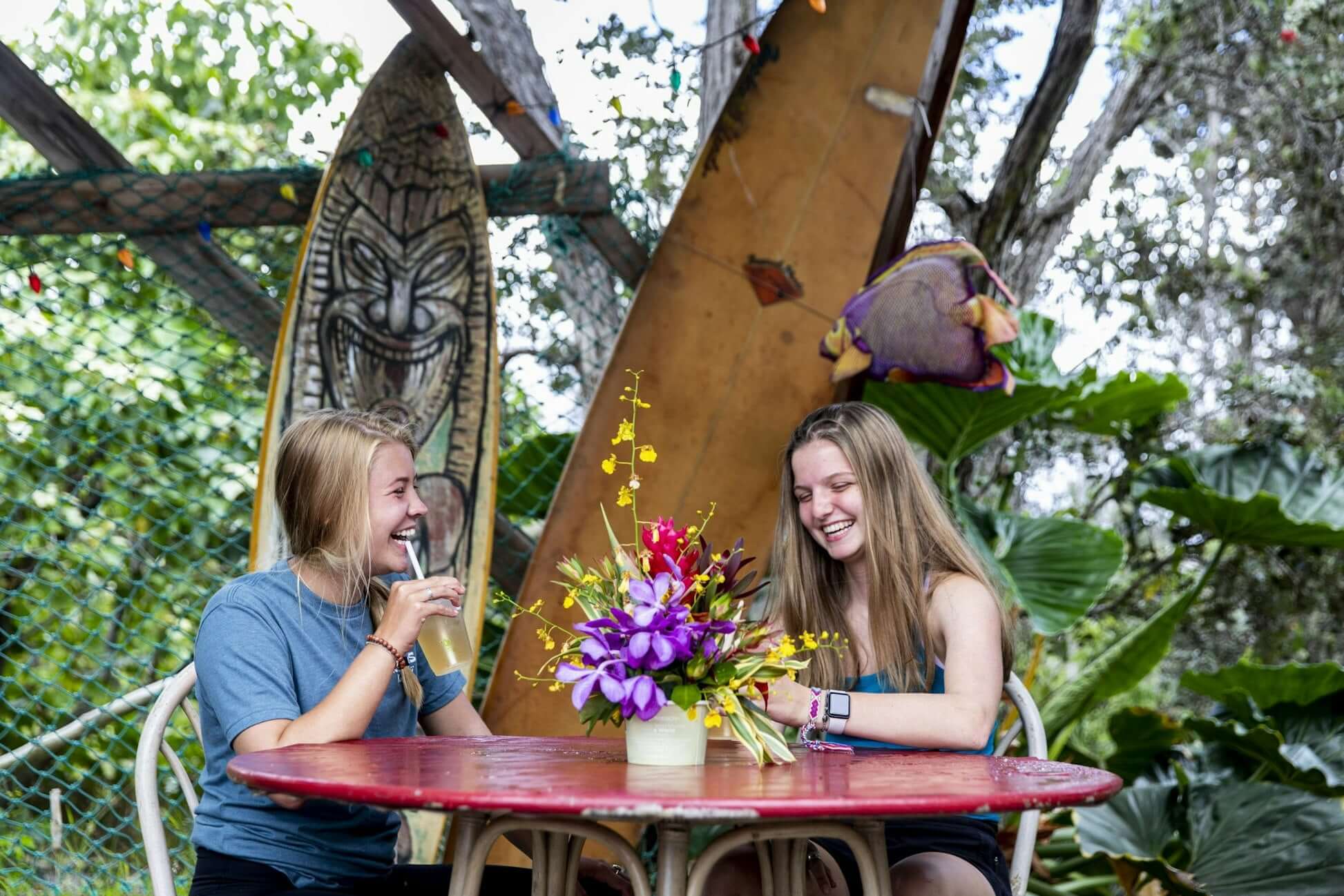 Two teens enjoying refreshments and laughter at a tropical table with a vibrant flower arrangement in Hawaii.