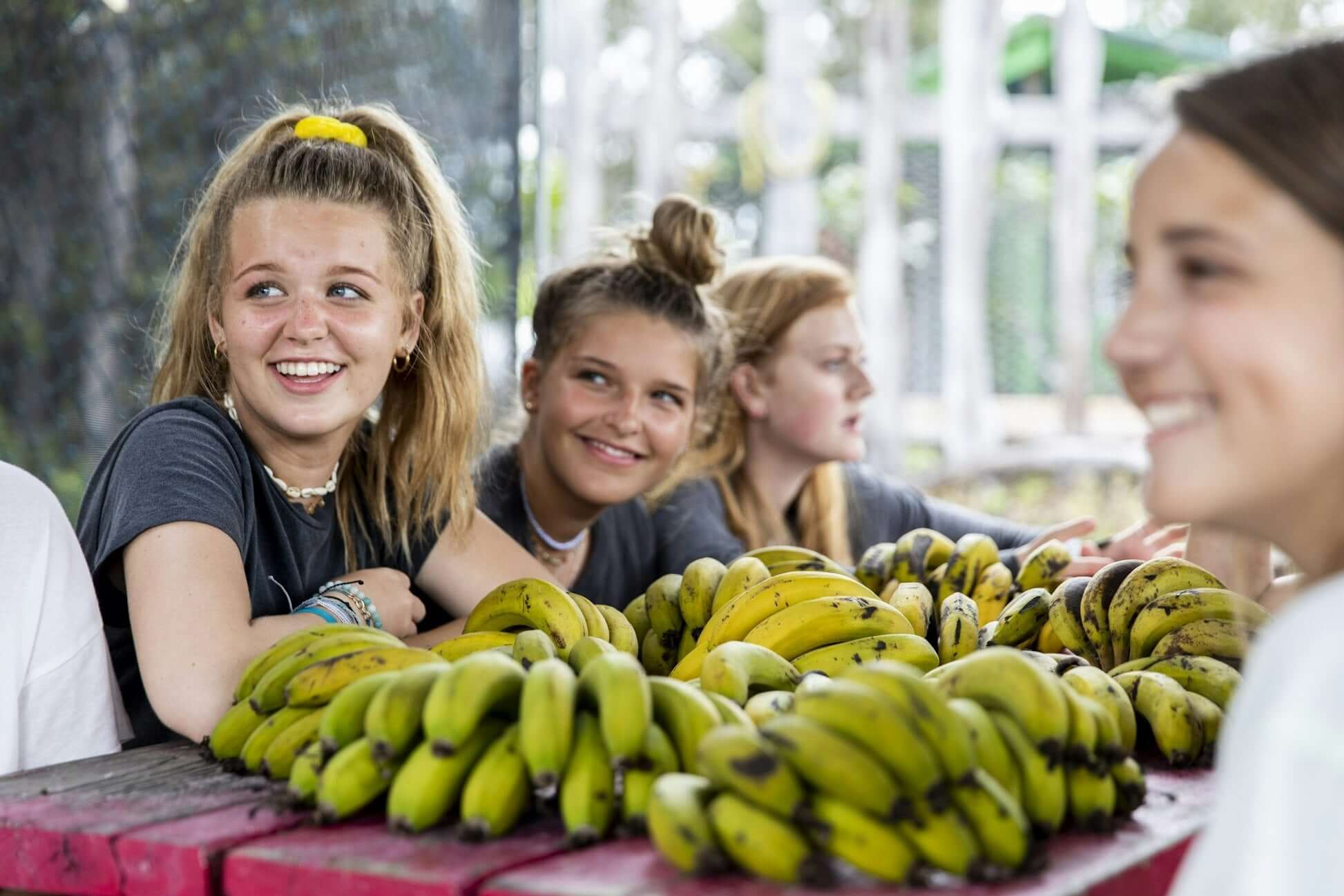 Teen girls smiling at a market table filled with fresh bananas, enjoying a community service experience in Hawaii.