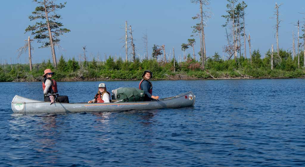 Three campers enjoying a canoe ride on a serene lake during a summer camp program, surrounded by lush greenery.