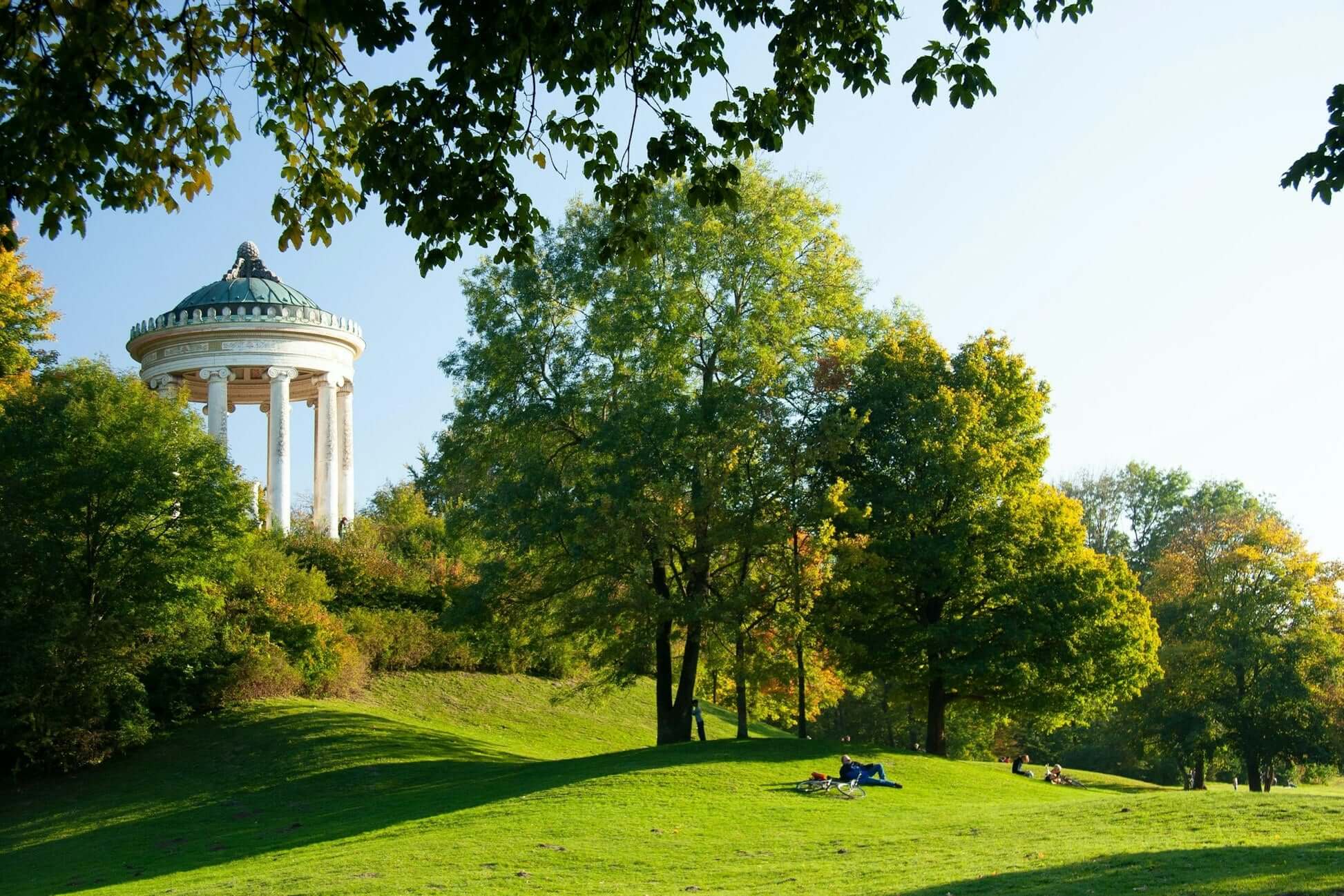 Sunny park scene in Munich with a historic pavilion and lush green trees, ideal for outdoor summer activities.