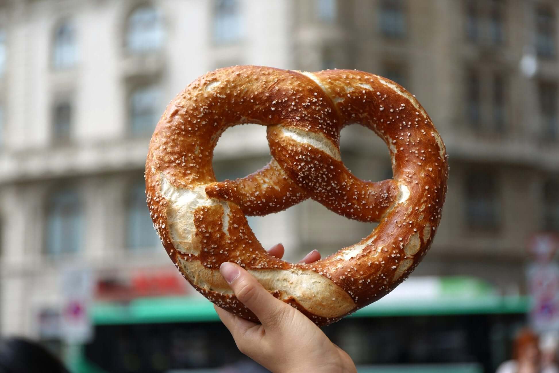 Hand holding a large, golden-brown pretzel sprinkled with salt against an urban backdrop.