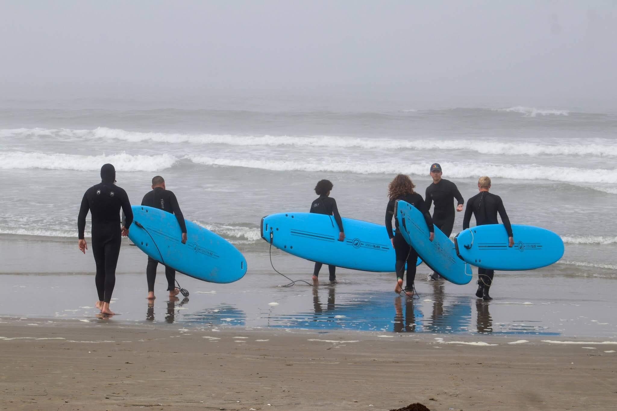 Kids and teens in wetsuits carrying blue surfboards along the beach at Camp Ocean Pines in California.