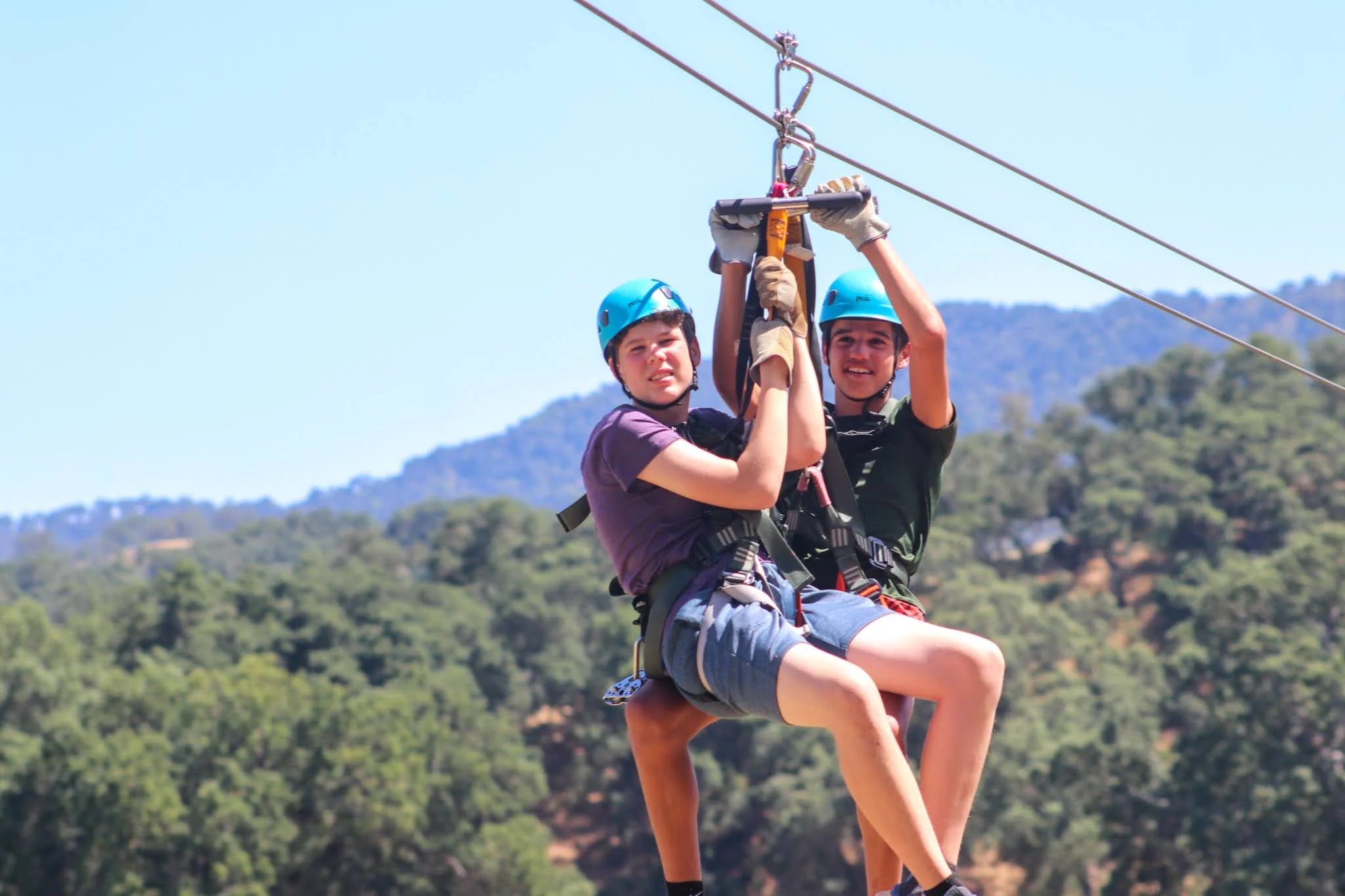 Two teenagers enjoying a zip line adventure at a summer camp, surrounded by a lush forest and blue sky.