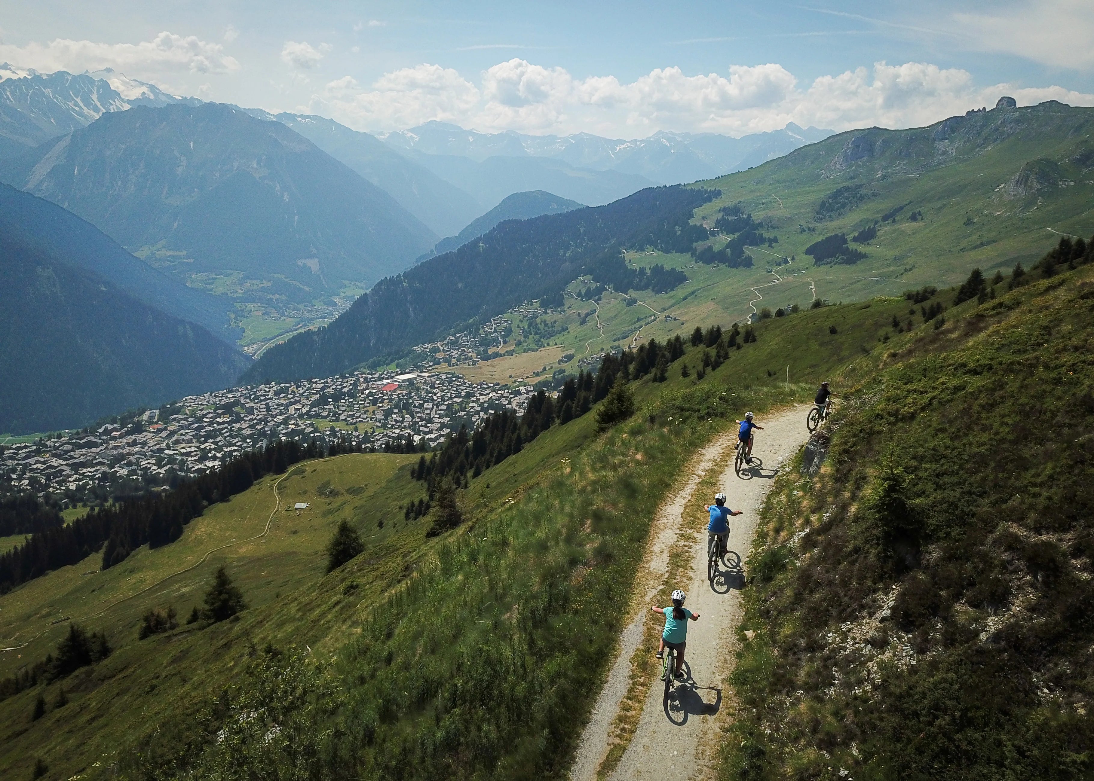 Aerial view of cyclists on a mountain trail surrounded by green hills and a town below, perfect for summer activities.