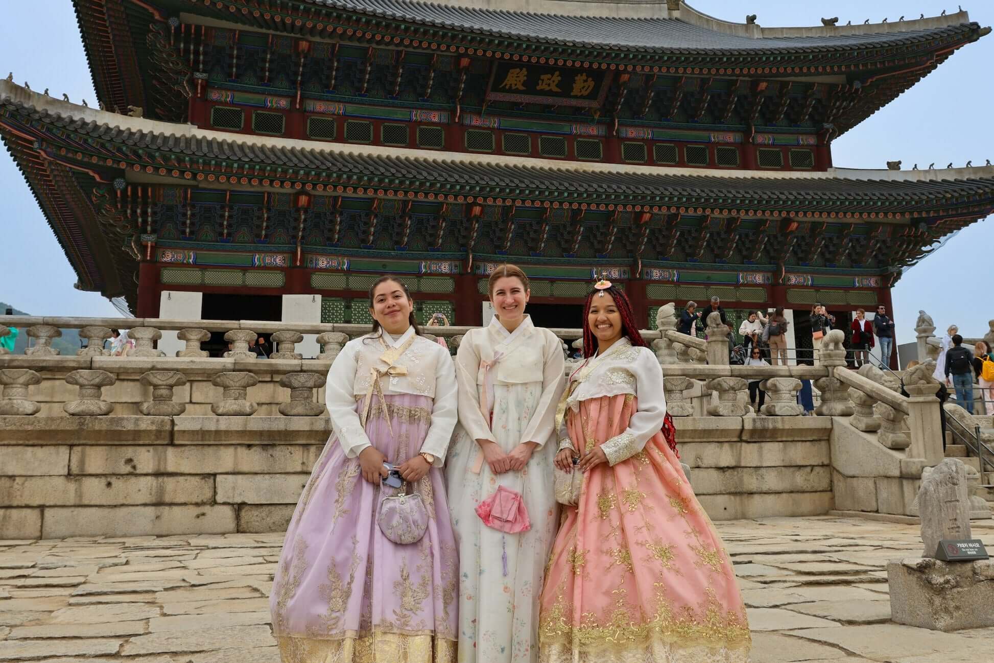 Three teens in traditional hanbok attire posing in front of a historic South Korean building.