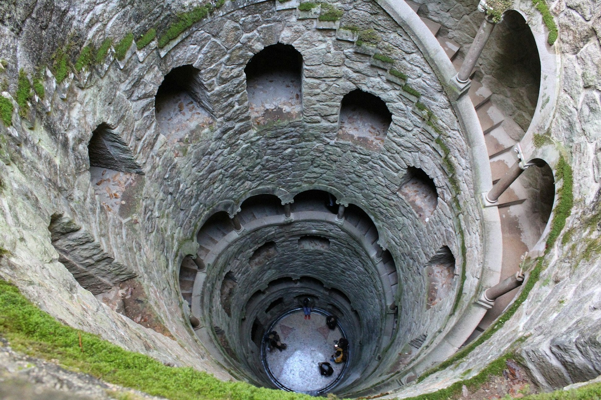 Aerial view of a moss-covered stone well with spiral stairs and visitors at the bottom, showcasing unique architecture.