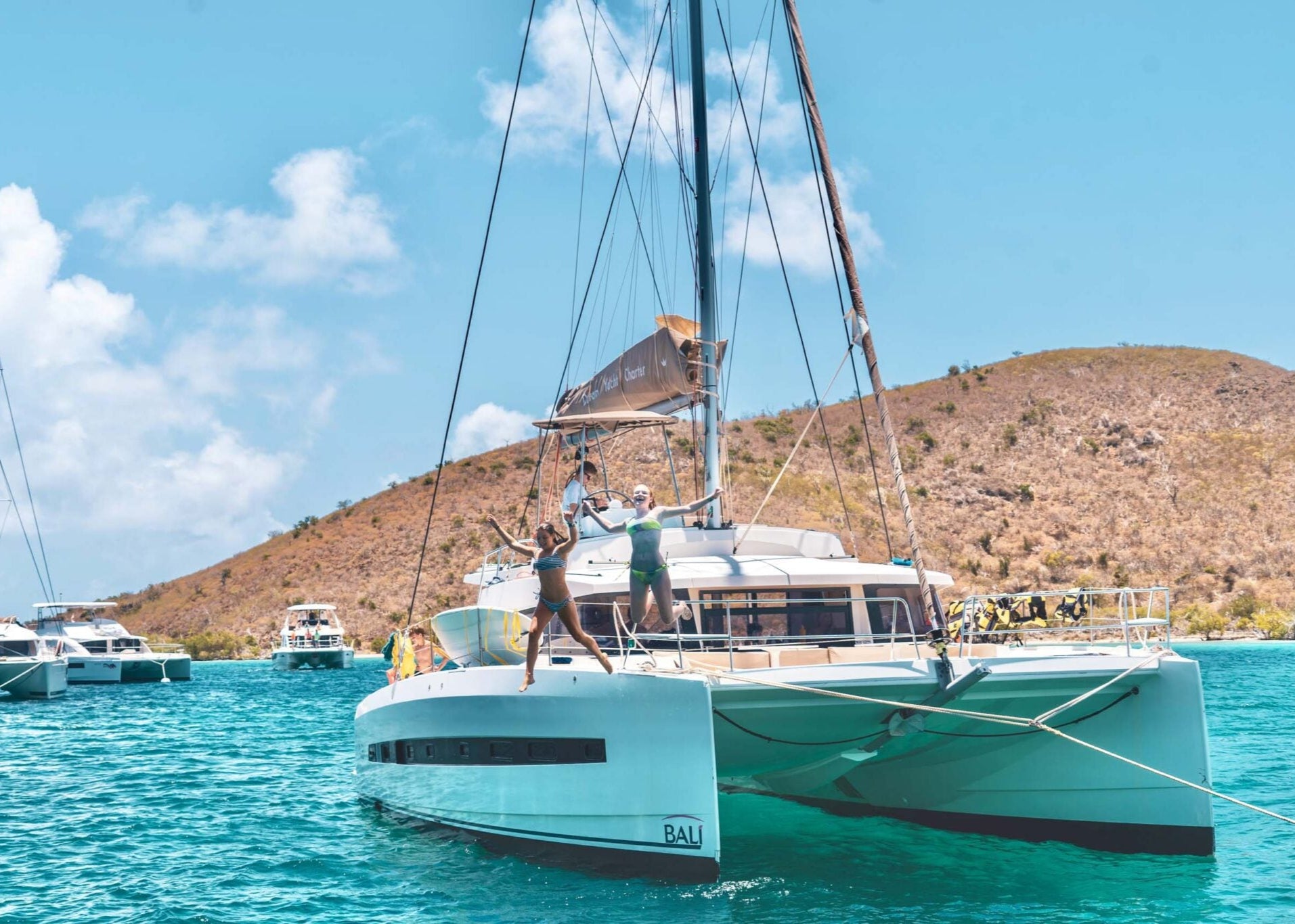 Catamaran sailing in turquoise waters with people on board, surrounded by boats and a scenic mountainous backdrop.