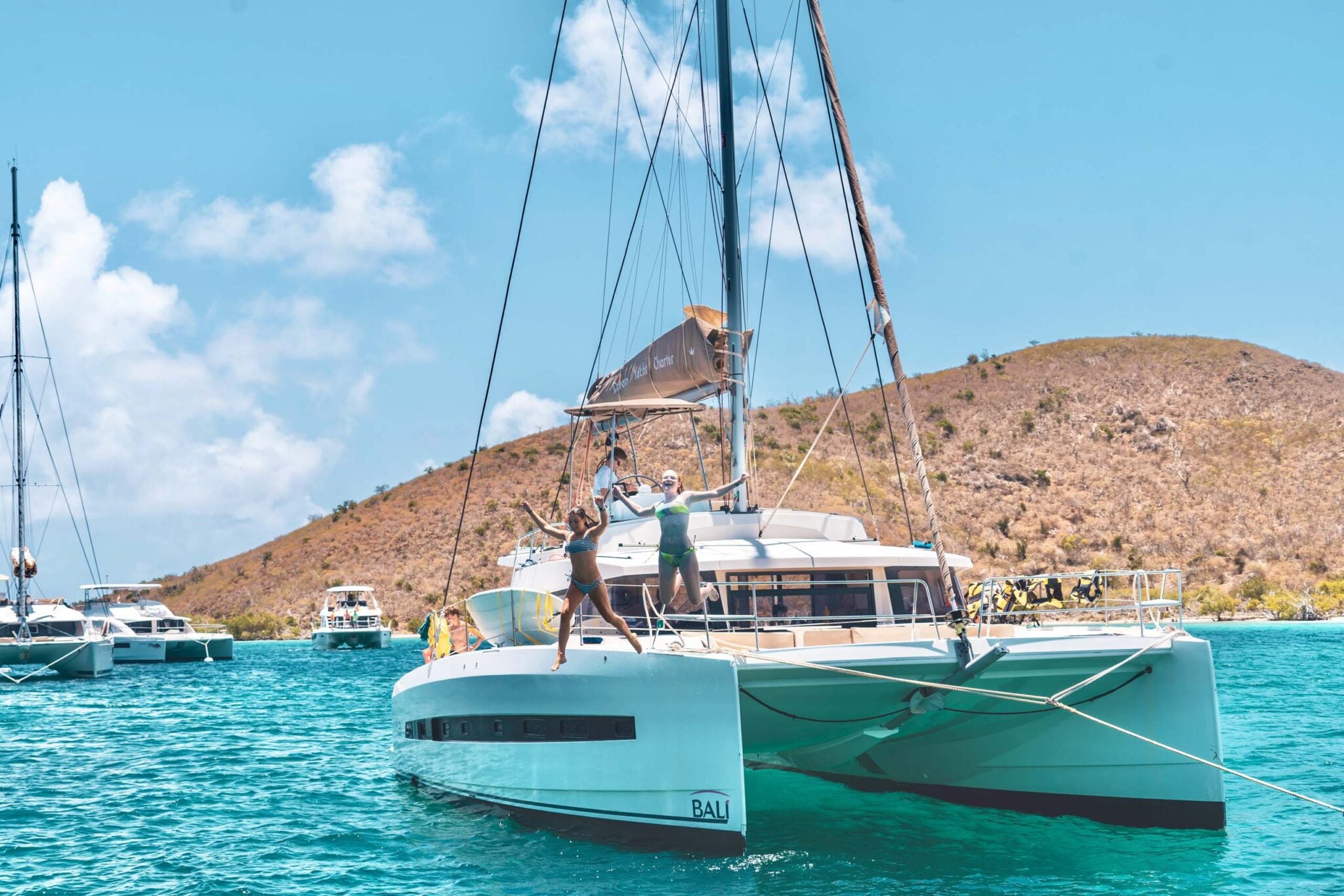 Catamaran sailing in turquoise waters with people on board, surrounded by boats and a scenic mountainous backdrop.