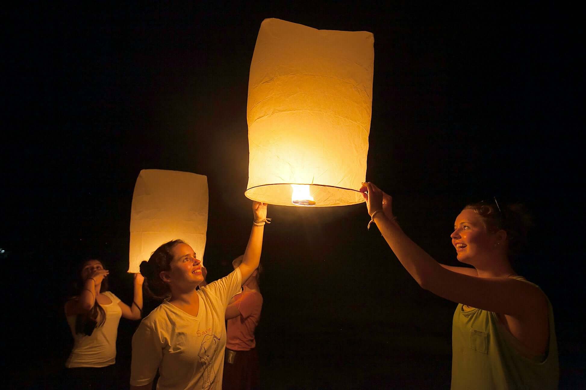 Teen travelers joyfully release glowing lanterns into the night sky, embracing Thai traditions during their adventure.