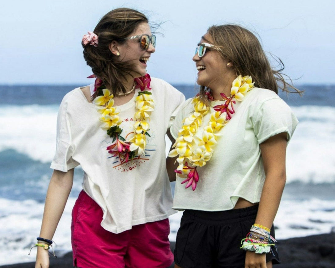 Two teens wearing leis smile at each other on a Hawaiian beach with ocean waves in the background, enjoying summer.