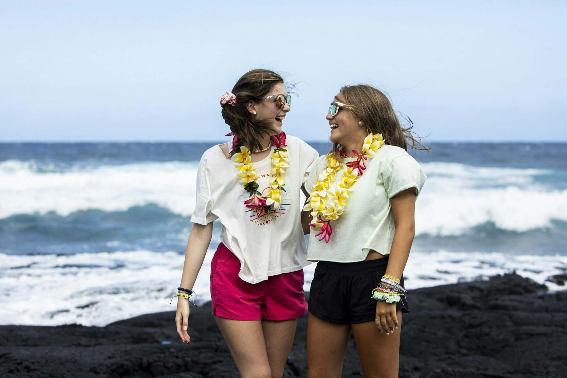 Two teens wearing leis smile at each other on a Hawaiian beach with ocean waves in the background, enjoying summer.
